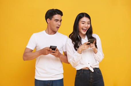 shocked-asian-man-spying-her-smiling-girlfriend-while-both-using-mobile-phones-isolated-yellow-backg (1)